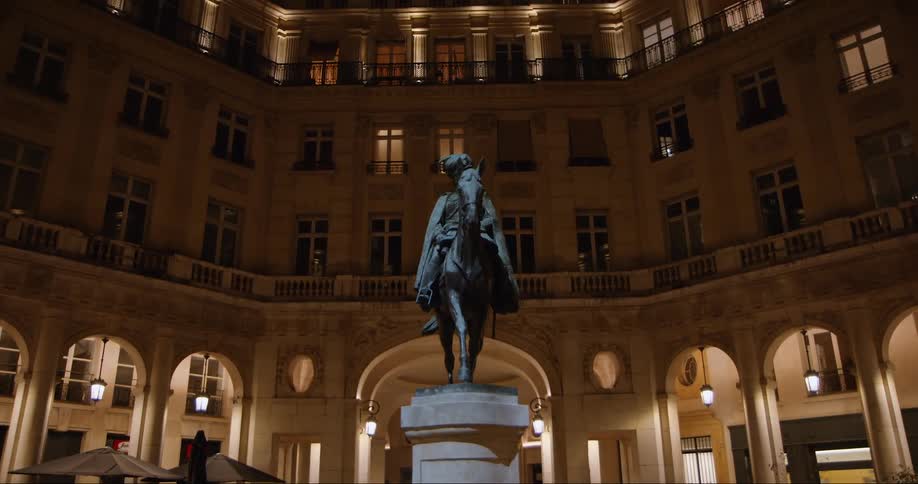 The camera moves in on a statue of a man sitting on a horse on a tall pedestal located in front likely in a courtyard of a tall multistory building with many windows some of which are lit others are dark