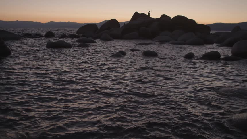 Someone standing on top of a rock formation as waves lap against the rocks during sunset