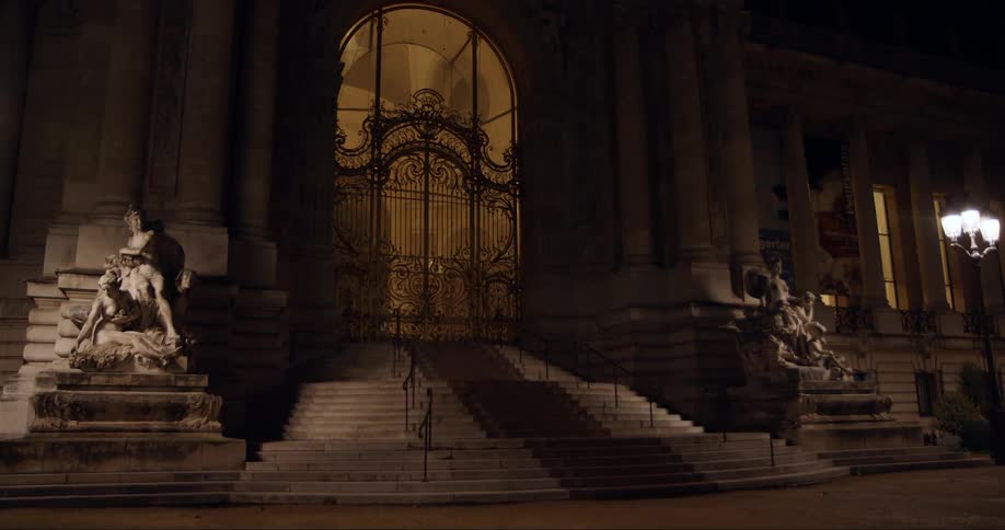 A large urban building with a big metal gate and large statues flanking stairs is lit up at night
