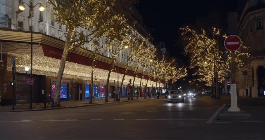 Cars travel down a city road in front of a litup building lined with trees