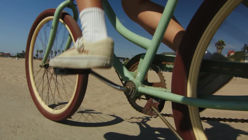 A closeup view of someone pedaling a bike over a path near sand and buildings