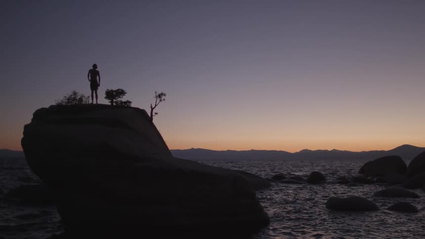 A person is standing on a large rock before the water at dawn