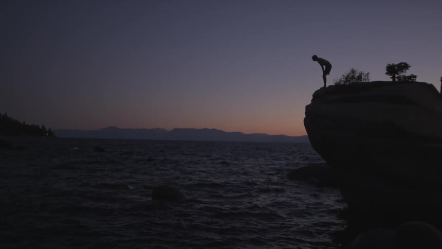 A person is standing on the edge of a cliff over water at dawn