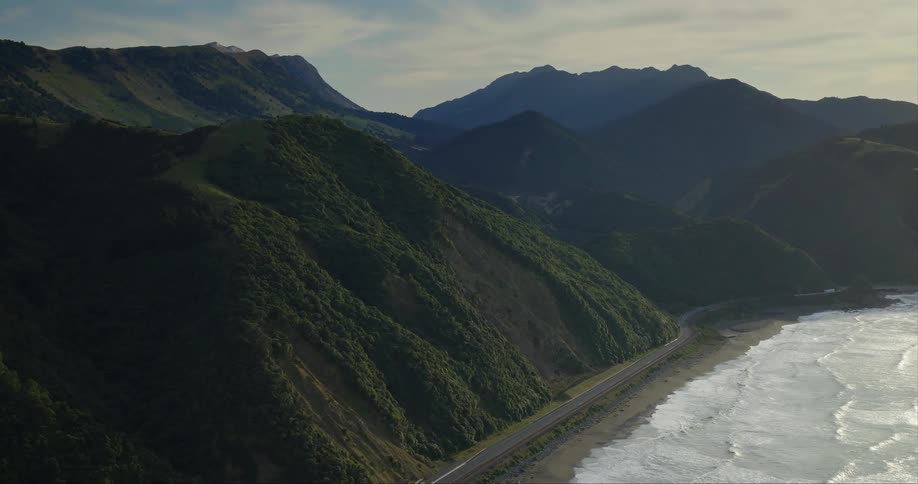 A landscape of a beach with crashing waves and several hills of green forests