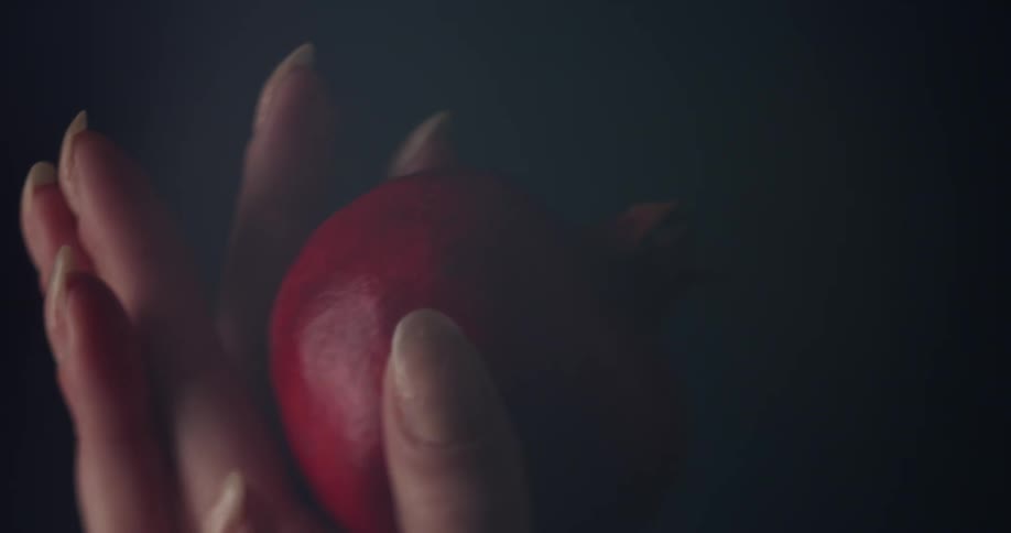Close up of a womans fingers wrapped around a small red fruit in a dark room
