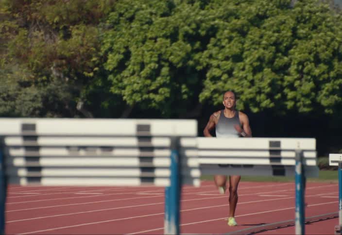 A woman runs on a track field next to hurdles