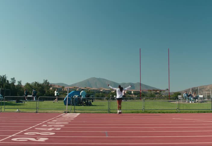 A female hops around on a running track with people in a field behind her