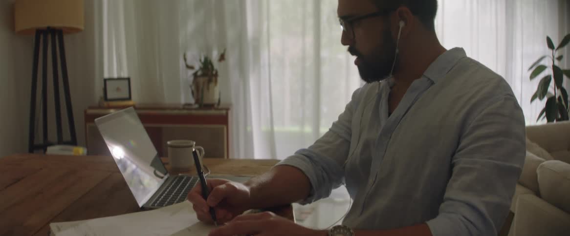 A man sitting at a table wearing headphones as he looks and nods at his laptop then writes on his notebook