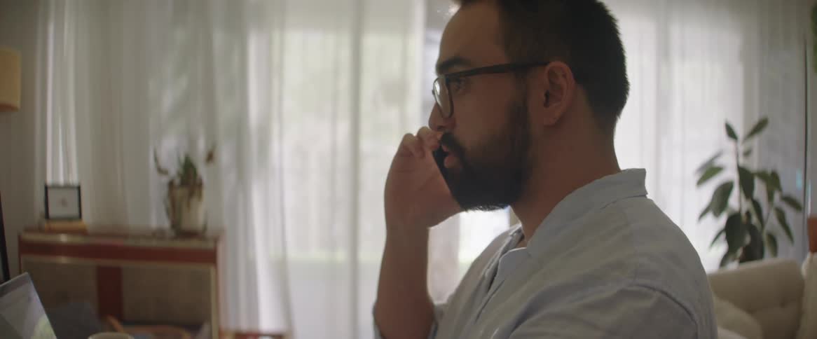 A man sitting is talking on the cell phone with someone and his hand is on top of a piece of paper with a pen on his finger