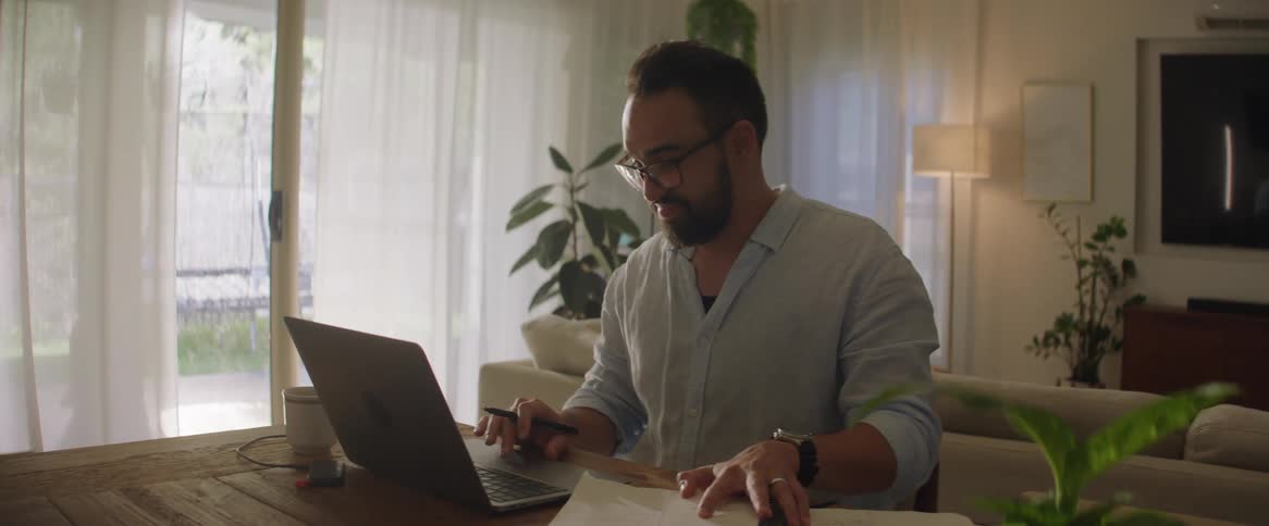 Man with beard glasses light blue shirt and wristwatch works at table typing on laptop