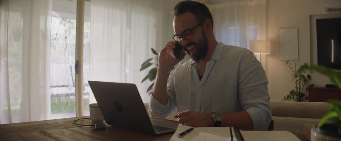 A man sitting at a table in his house talking on his cellphone in front of his laptop