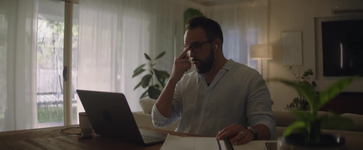 A man is sitting at a table in front of a laptop computer talking with a pen in his hand and open notebooks