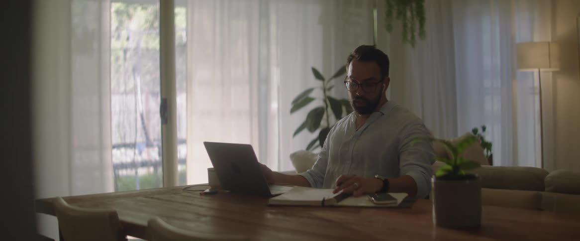 A man is sitting at a table in front of a laptop computer talking with a pen in his hand and open notebooks