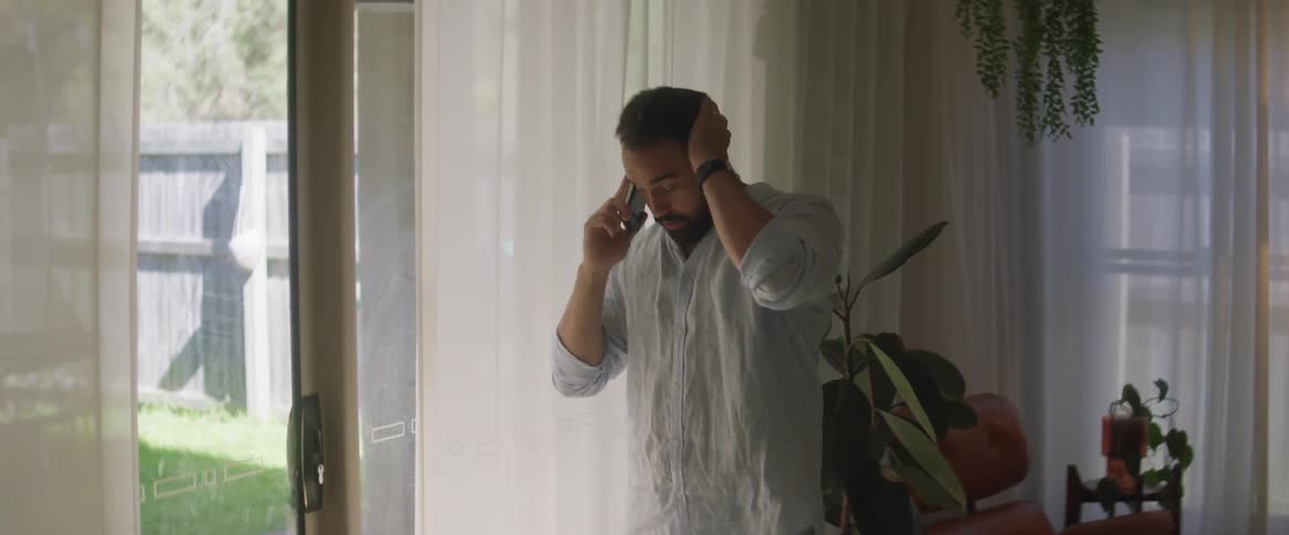 A man is standing in a room near windows talking on the phone and rubbing his face and head as he speaks