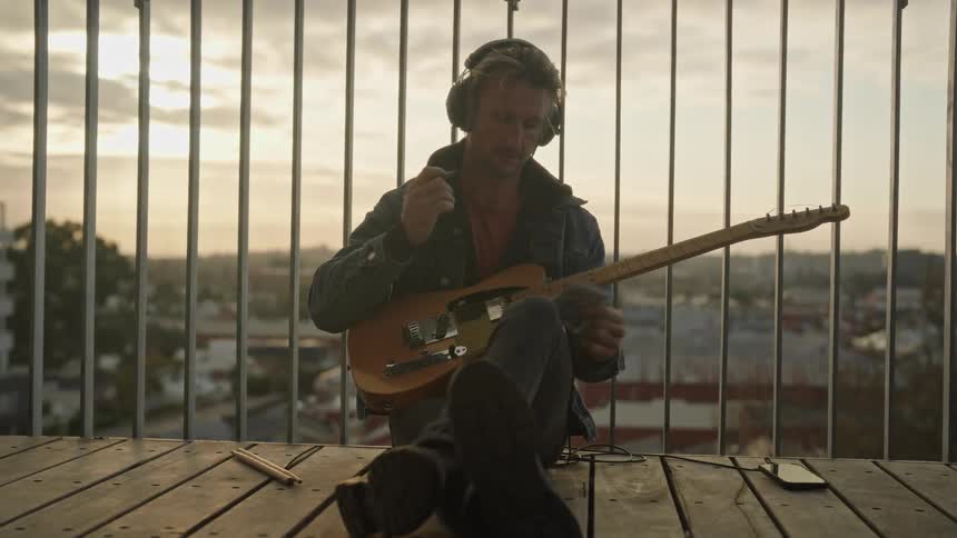 A man sitting with headphones and his guitar making notes in his notebook