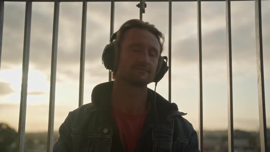 Man wearing headphones reclines on a fence at sunset