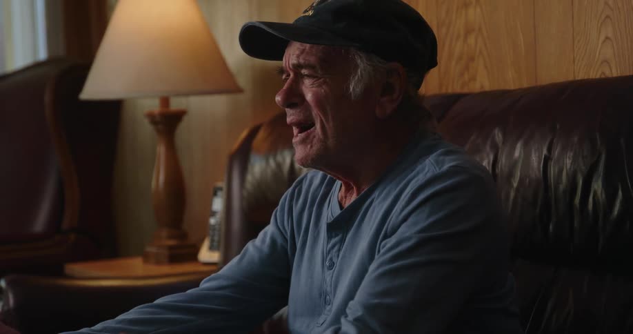 A man sitting on his leather couch wearing a baseball cap as he raises his arms and yells in frustration