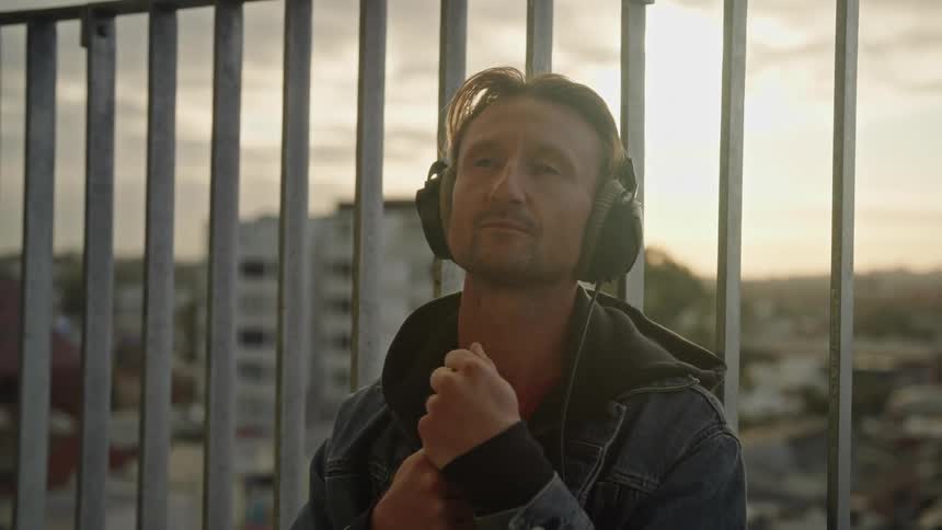 A man standing outdoors with headphones on holding a drum stick looking contemplatively as the sun sets on the horizon behind him