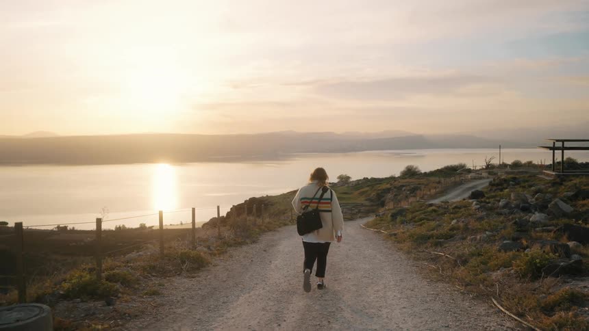 The backside of a woman wearing a white sweater and black bag as she walks on a coastal road as the sun sets on the horizon