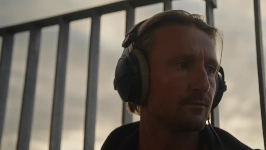 A man on a balcony wearing large headphones uses drumsticks to keep up with the music hes listening to