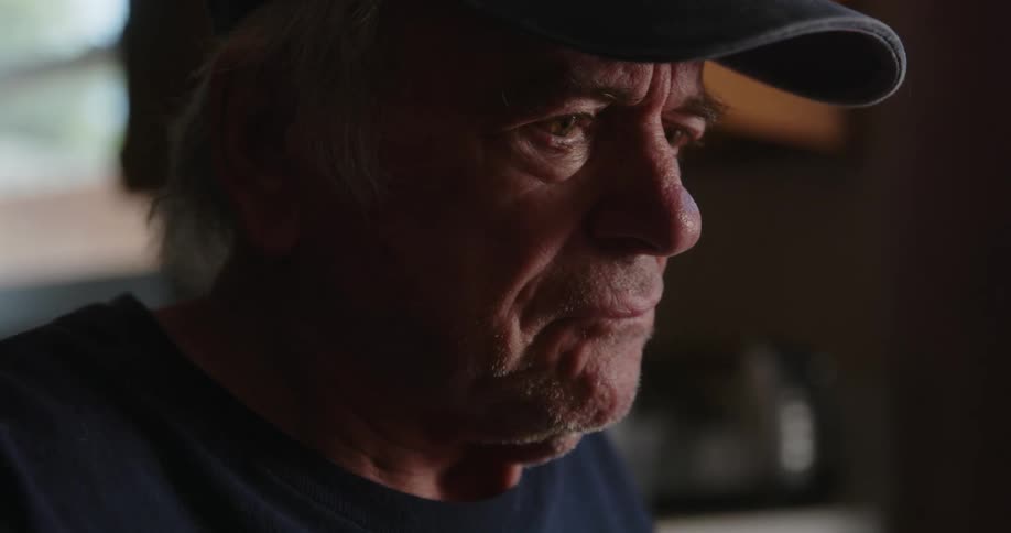 An older mans face wearing a baseball cap as he looks forward with a furrowed brow
