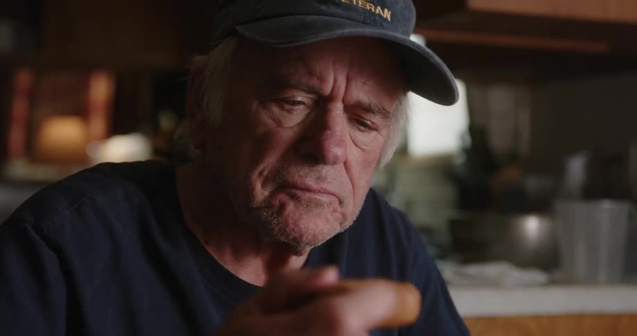 An older man sitting in his home wearing a baseball cap and tshirt eating food with his hands while watching something in front of him
