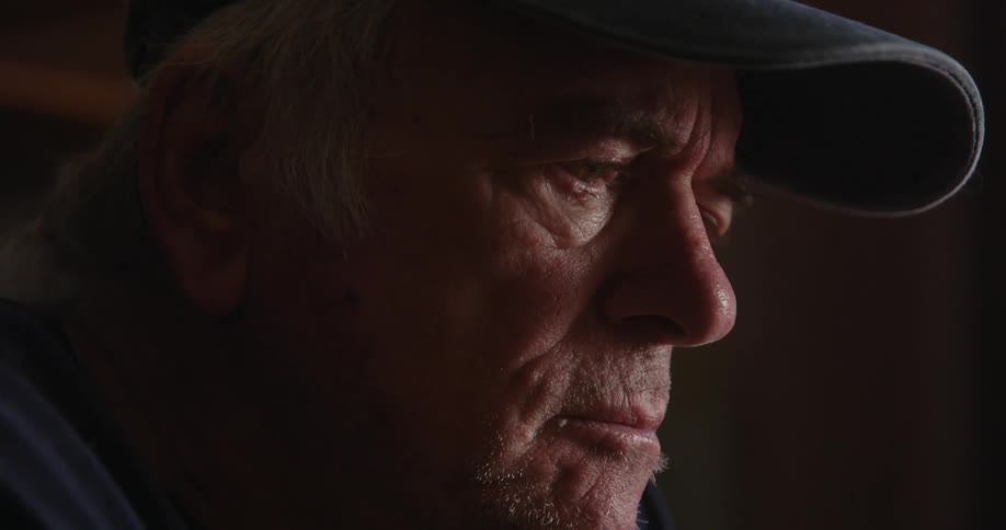 An older mans face as he eats food with his fingers and scoffs at something he is watching