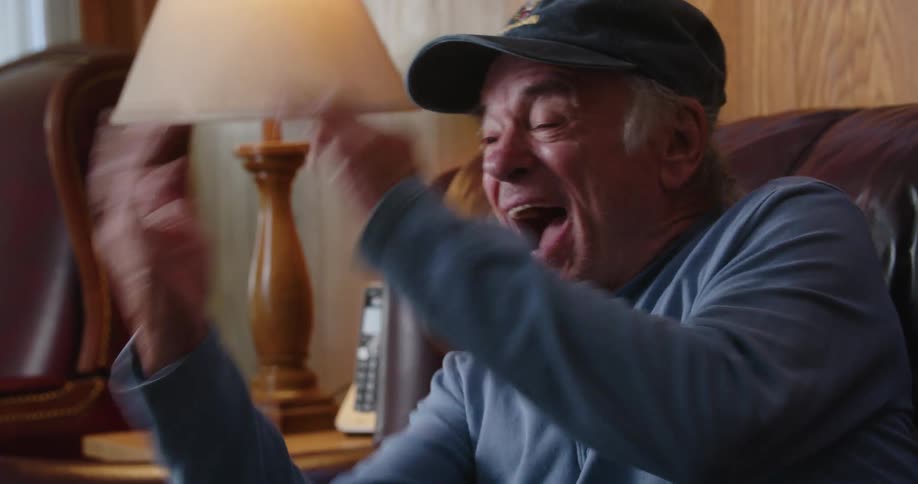 An older man sitting on his couch cheering and smiling as he raises his arms and hands in excitement