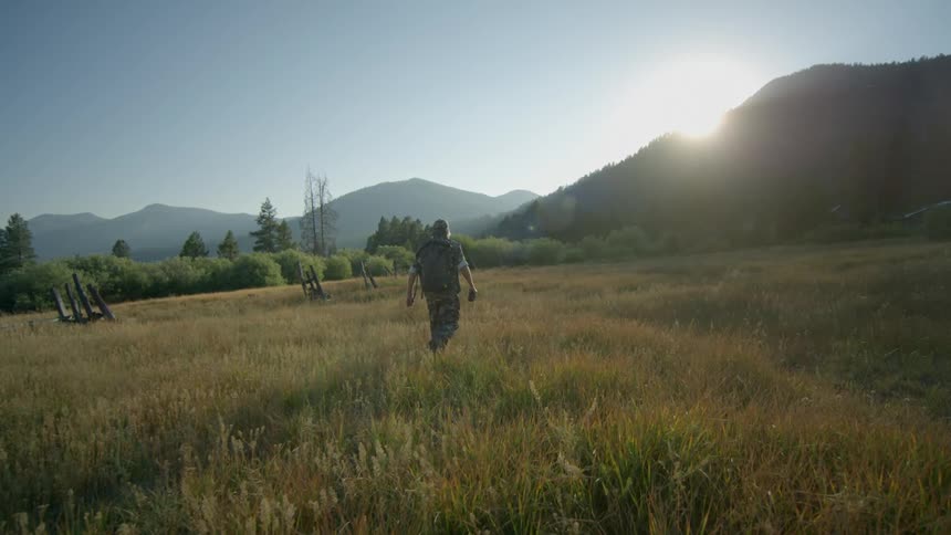 A man dressed in camouflage walks through a field and heads towards the mountains