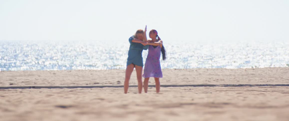 Two women perform dance routine on beach as waves sparkle in sun