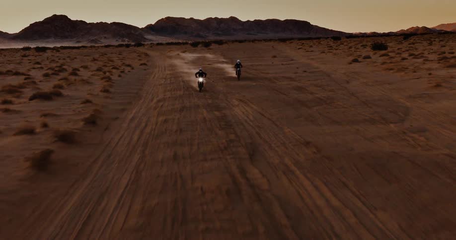 Two people ride motorcycles across a flat desert land at sunset