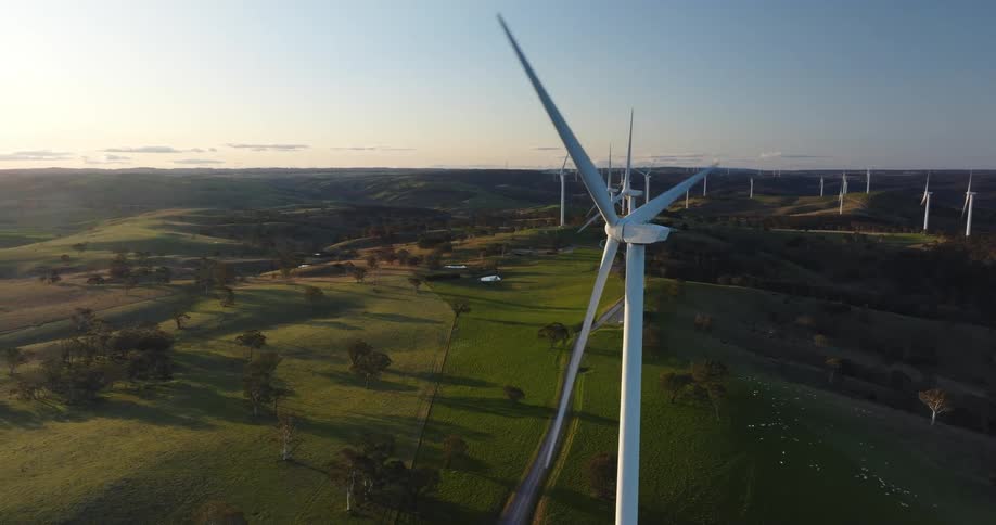The sun shines upon wind turbines as they spin on a wind farm along a rural road