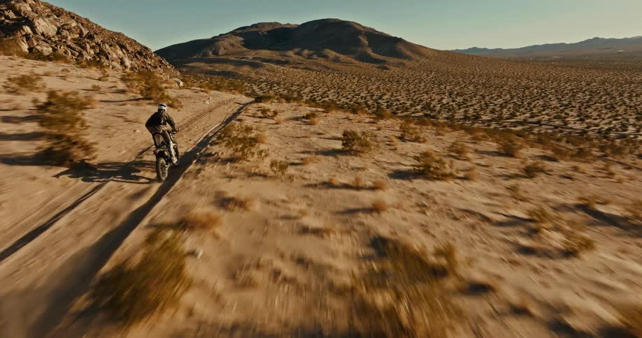 The camera chases a man racing across uneven terrain in the desert on a dirt bike