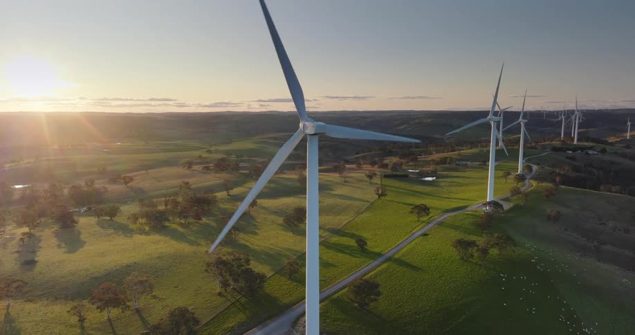 The sun shines brightly as wind turbines spin on a wind farm along a rural road