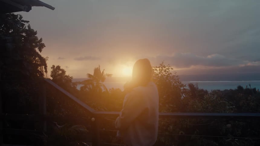 A woman on a balcony holding a baby and staring at the sunset