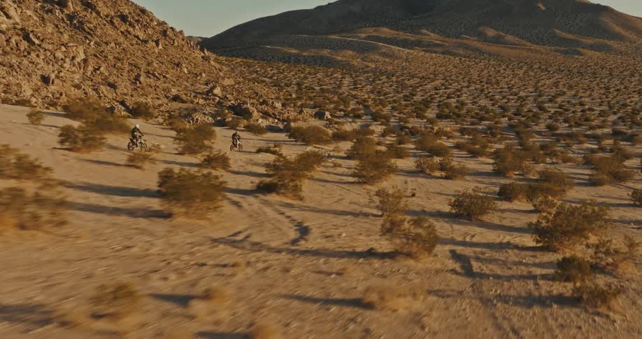 The camera zooms across the desert as two friends chase each other through the dunes on dirt bikes
