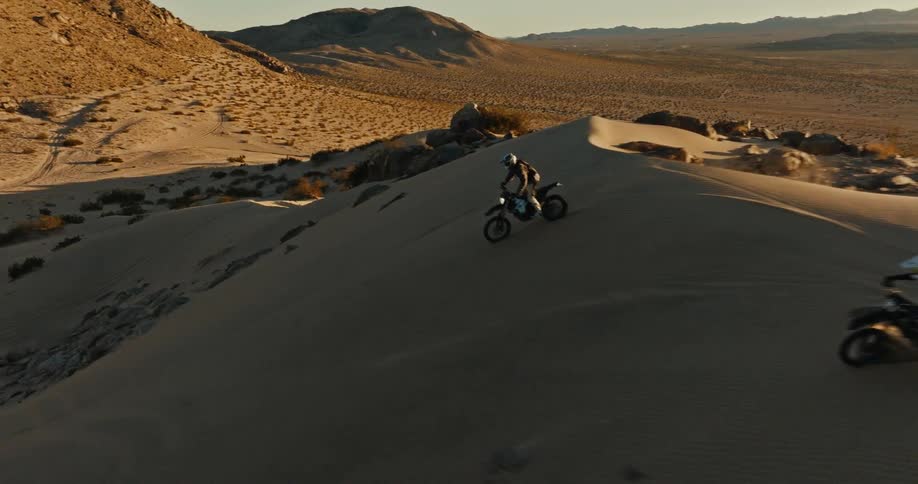 Motorcyclists ride along desert slopes with scrub land and mountains in the background