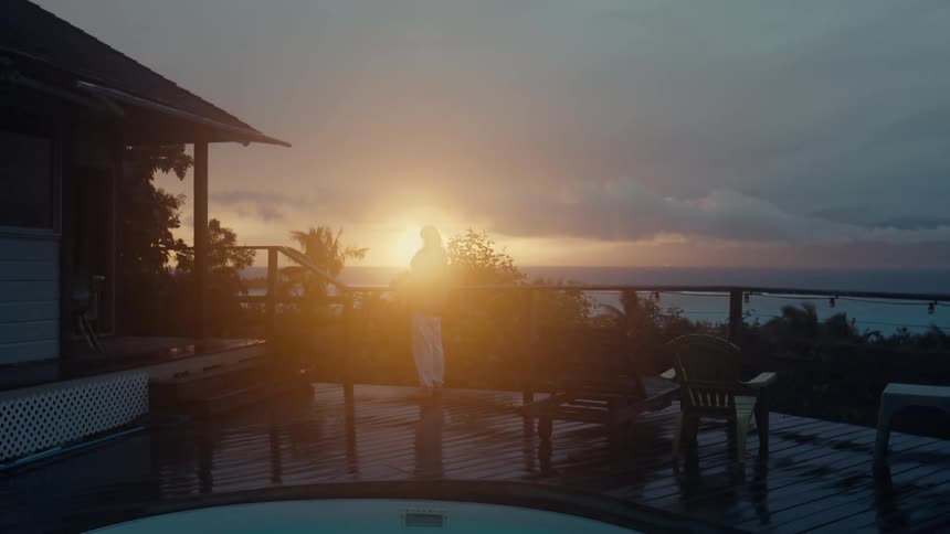 Woman stands on deck near pool looking out to the bright setting sun over the ocean on an overcast day