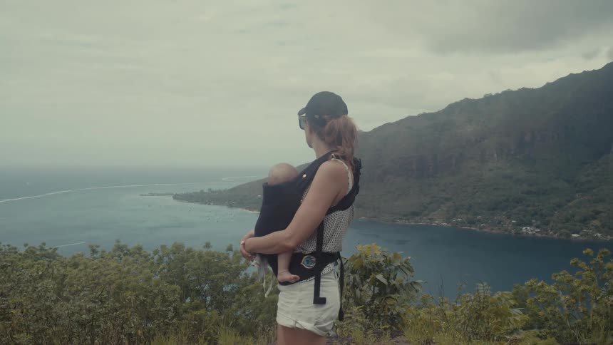 A mother holds her baby while overlooking the ocean coastline from a high vista