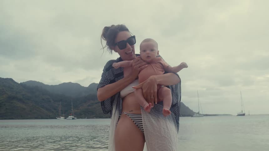 Mother holds baby while standing on beach with sailboats in the background on an overcast day