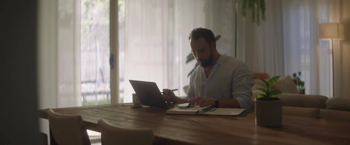Man working at home uses smartphone for calculations while viewing laptop