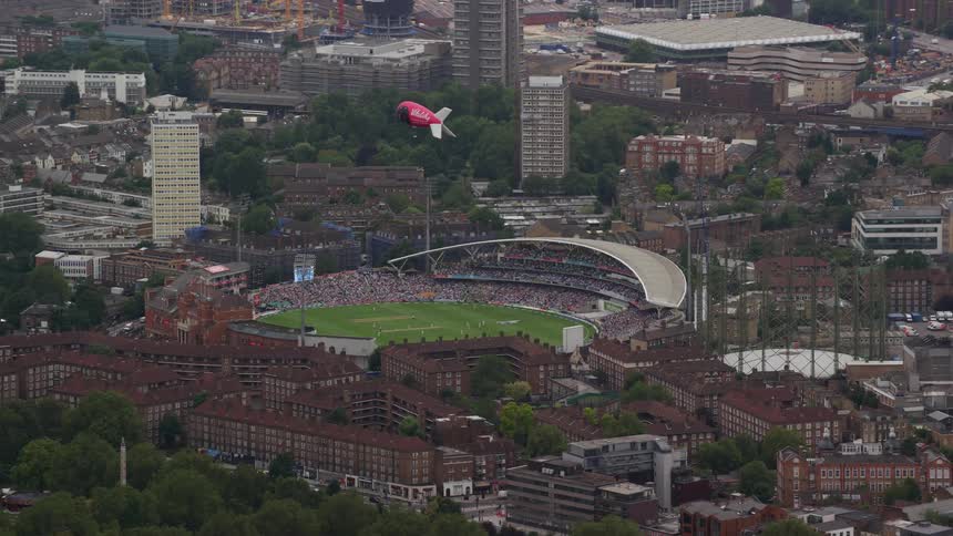 A sports arena with green grass next to residential buildings and a red airship in the sky