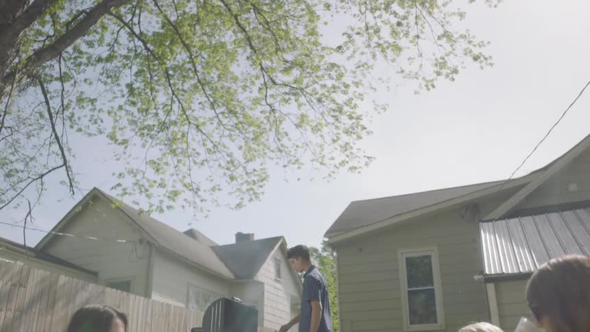 People sit in a yard talking as a person cooks on a grill