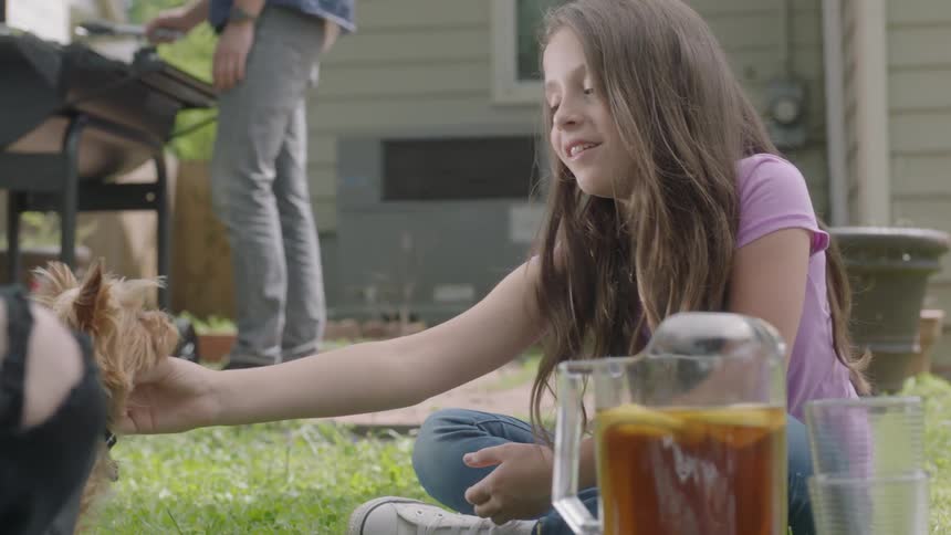 A girl is sitting in the grass petting a dog as a man is cooking on a barbecue