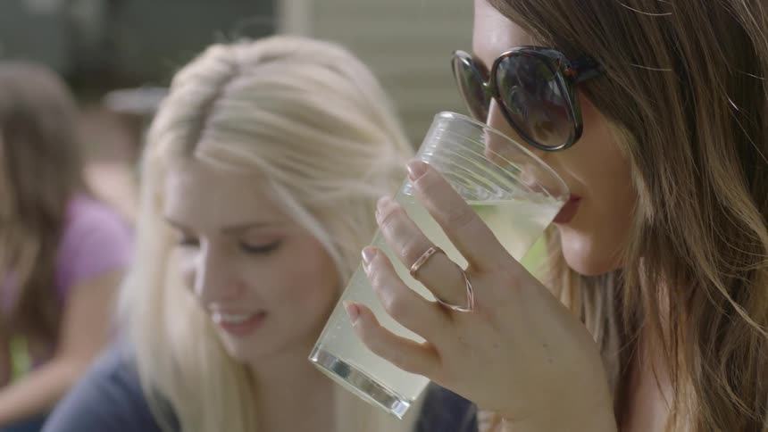 A woman picks up a glass and takes a drink as another eats fruit