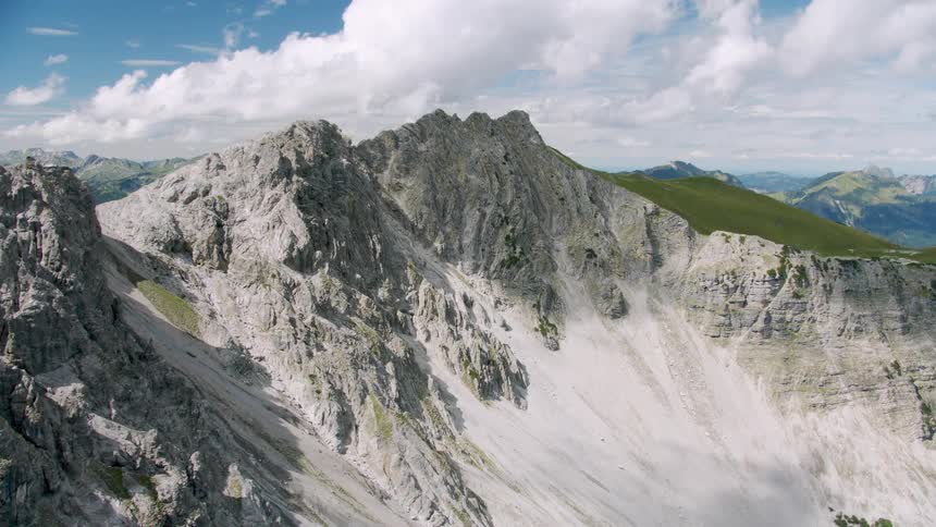 The sun shines upon a rough mountain ridge with mountains in the distance under a cloudy sky