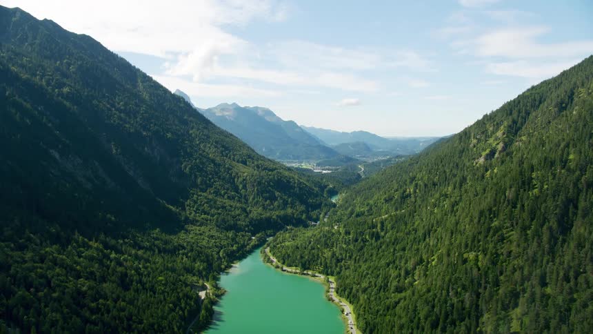 Traffic drives along road following shore of river at base of forest covered mountains