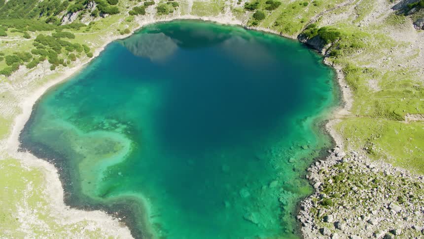 A mountain range and a clear lake of a surreal unnatural bluegreen color surrounded by rocky canyon walls with some shrubs and greenery amidst the rocks