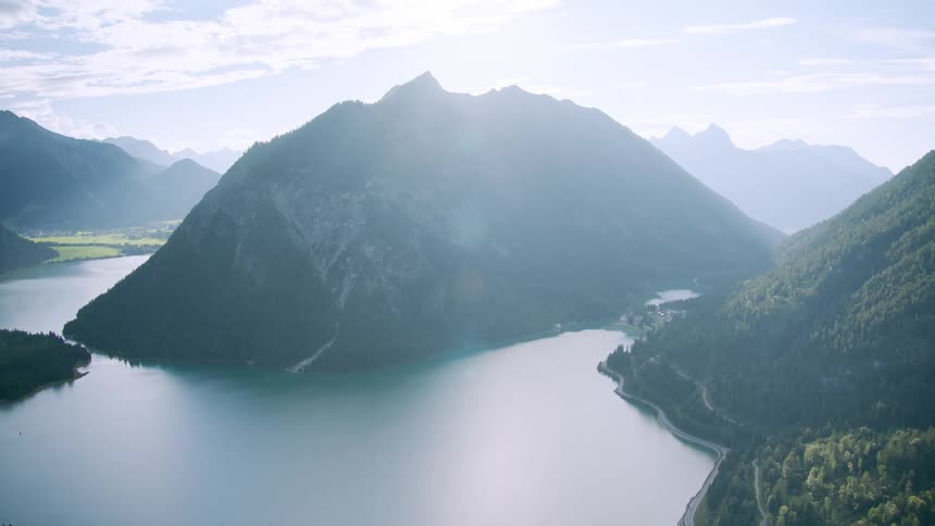 The sun shines brightly upon a lake surrounded by forested mountains under a cloudy hazy sky