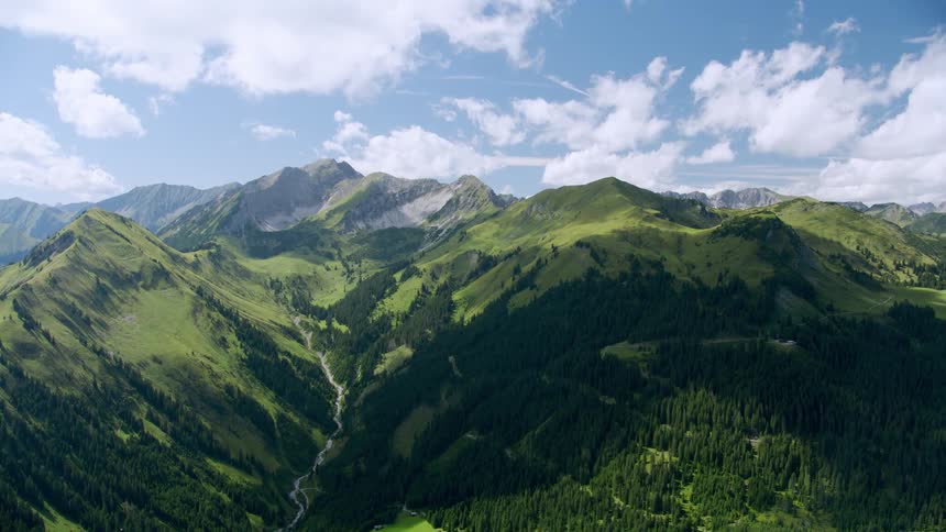 Mountain range with green trees on the slopes at least below the treeline and there is a valley with a river beneath a blue sky with clouds